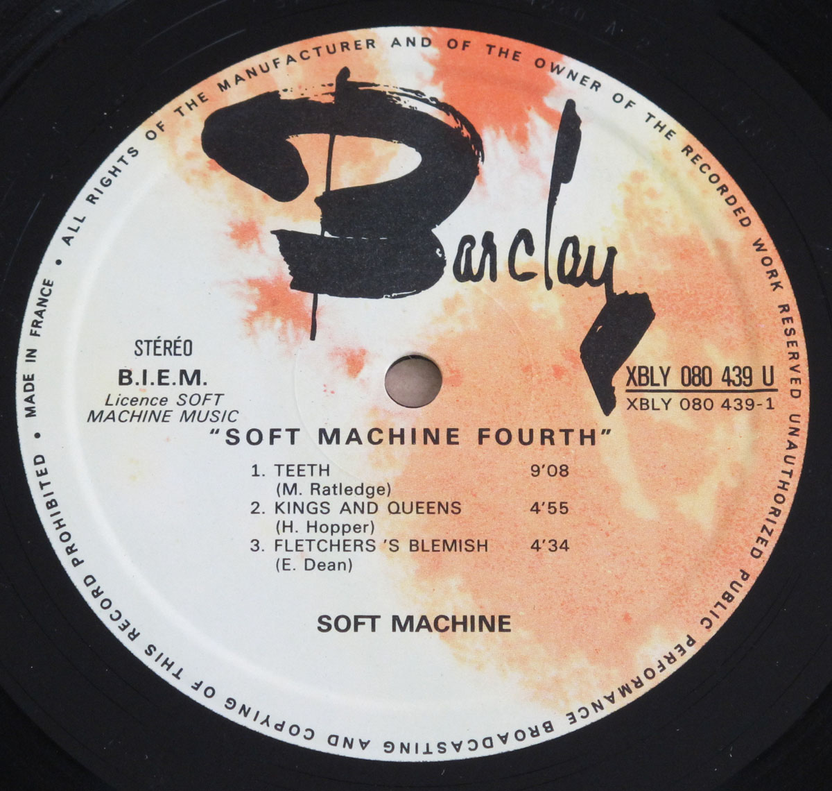 Close-up of the Side One label of Soft Machine's album 'Fourth' released on Barclay Records. The label is white with an orange watercolor-like splash effect and features the bold, black 'Barclay' logo in a hand-drawn style at the top. It lists three tracks: 'Teeth,' 'Kings and Queens,' and 'Fletcher’s Blemish,' along with their respective composers and durations. The catalog number 'XBLY 080 439 U' is printed on the right side.